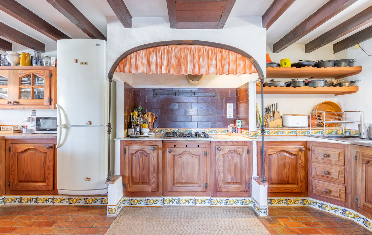 Cuisine de style traditionnel valencien avec poutres en bois apparentes, sol en terre cuite et zone de cuisson encadrée par une arche décorative carrelée.