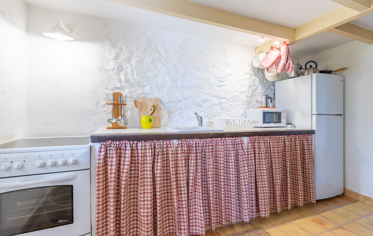 Cuisine rustique avec murs en pierre blanchie à la chaux, poutres apparentes et sol en terre cuite. Comprend des appareils blancs et un plan de travail avec rideaux à carreaux.