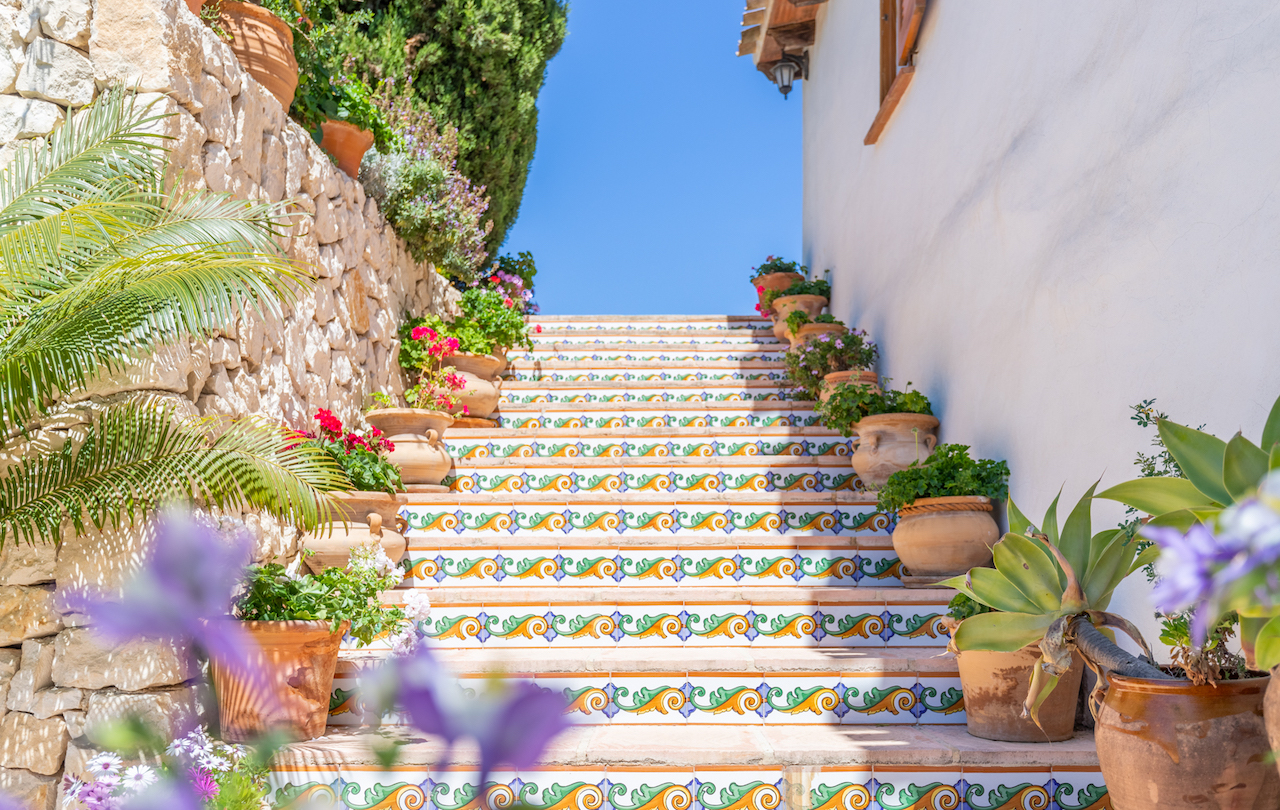 Escalier extérieur orné de contremarches en azulejos traditionnels, bordé de pots en terre cuite fleuris et d'un mur en pierre sèche typique.