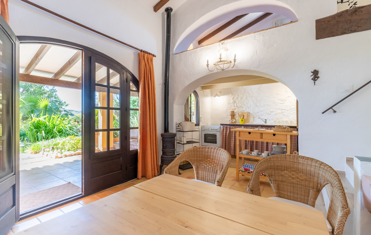 Espace salle à manger rustique avec poutres en bois, poêle en fonte et sol en terre cuite. Une arche traditionnelle s'ouvre sur une kitchenette avec accès direct à la terrasse.