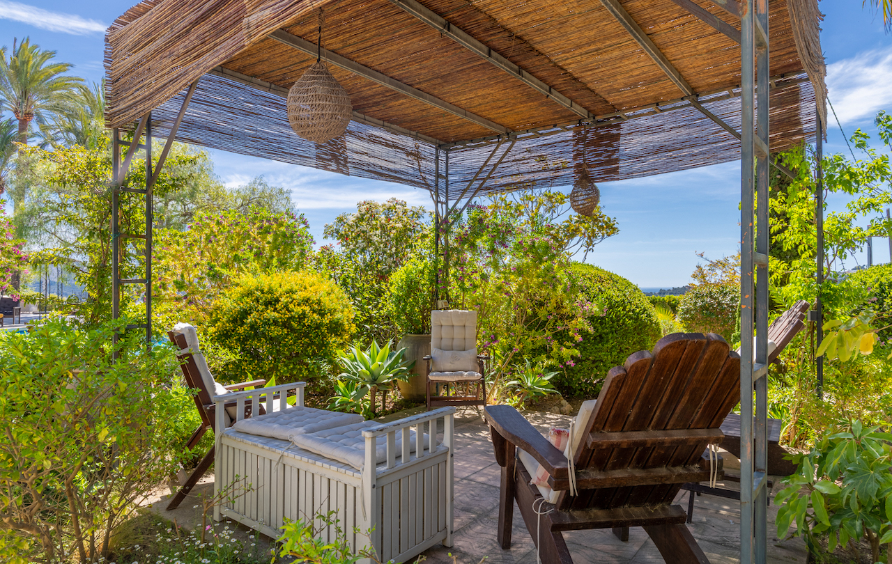 Pergola à structure métallique et toit en canisse abritant un coin salon, entourée d'un jardin méditerranéen avec vue sur la mer.