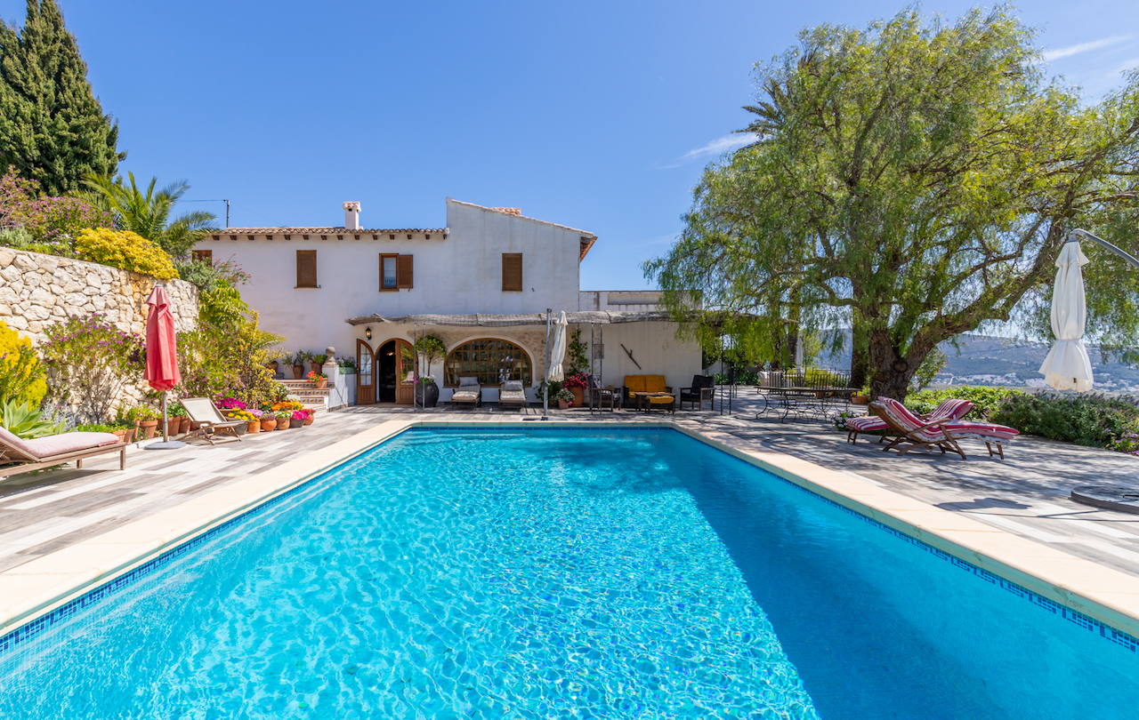 Piscine rectangulaire entourée d'une vaste terrasse en grès antidérapant, face à une façade traditionnelle blanche et un jardin arboré avec des murs en pierre.