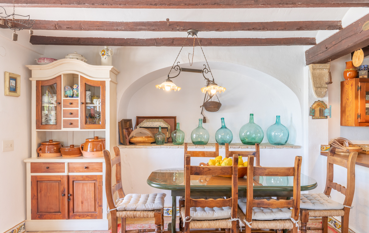 Salle à manger rustique valencienne avec poutres apparentes, buffet traditionnel et niche décorative ornée de dames-jeannes en verre derrière la table.