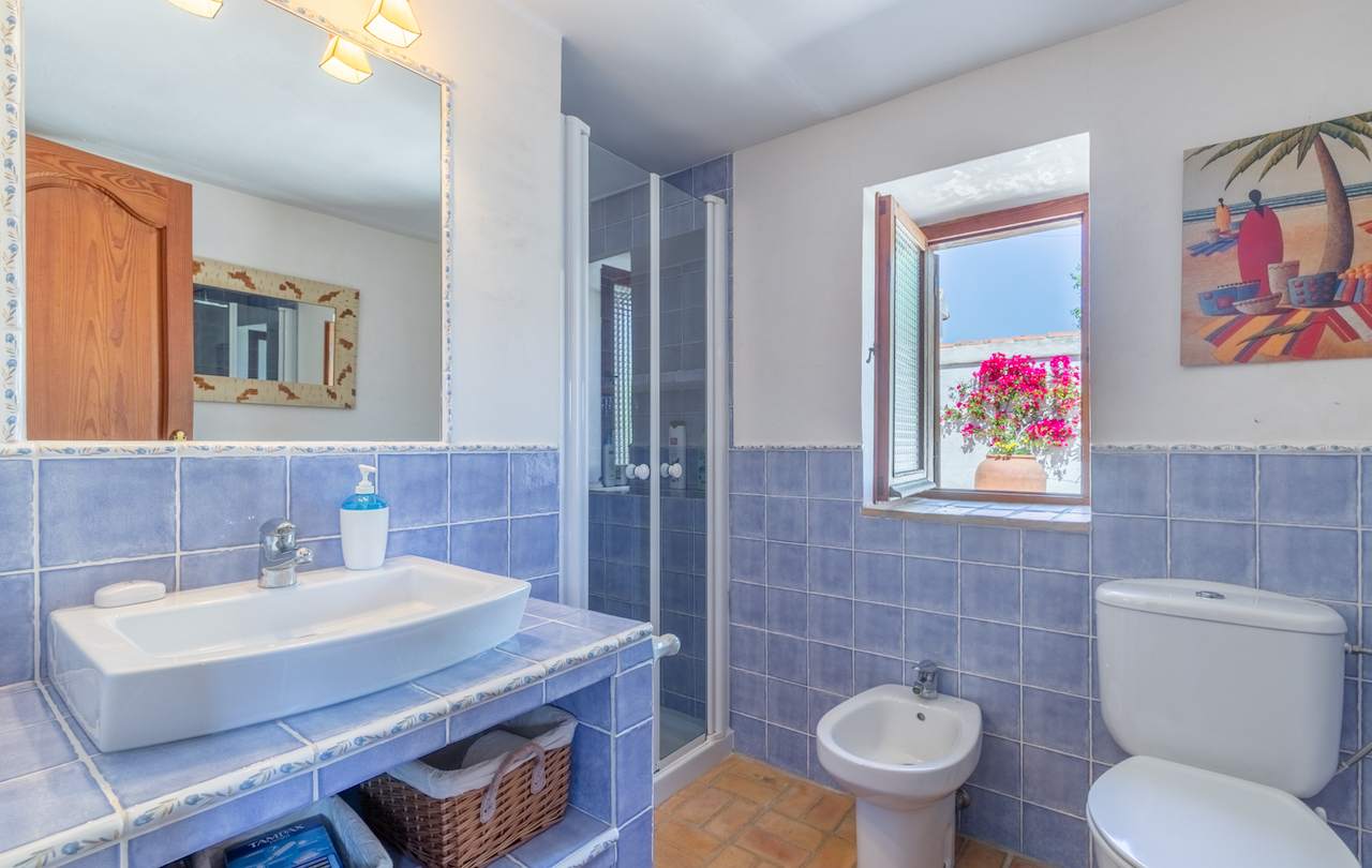 Salle de bain traditionnelle avec carrelage bleu, sol en terre cuite et fenêtre en bois sur le patio. Elle comprend un lavabo intégré, un bidet et une douche vitrée.