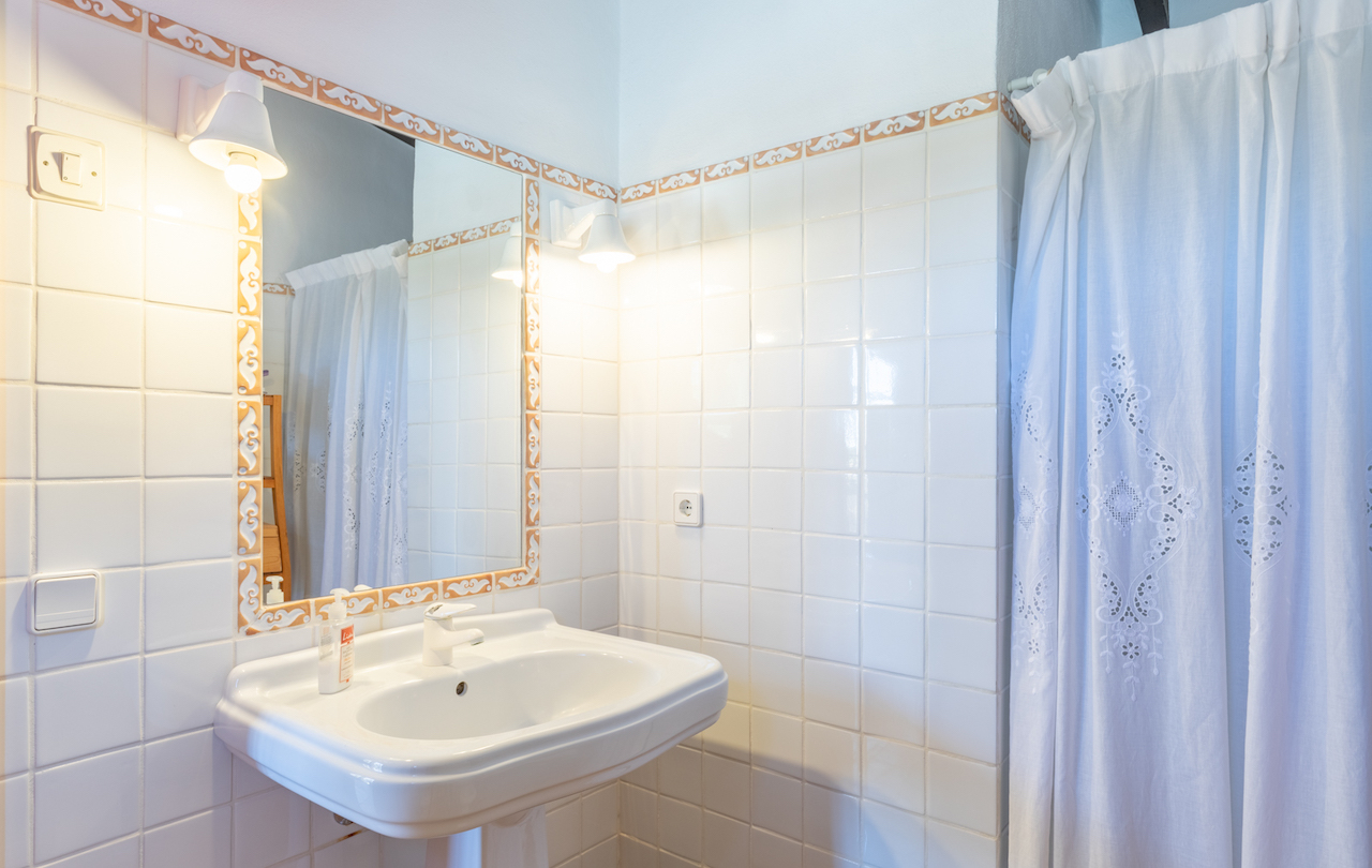 Salle de bain traditionnelle avec lavabo sur colonne, miroir encadré de carreaux décoratifs et appliques murales classiques près d'un rideau brodé.