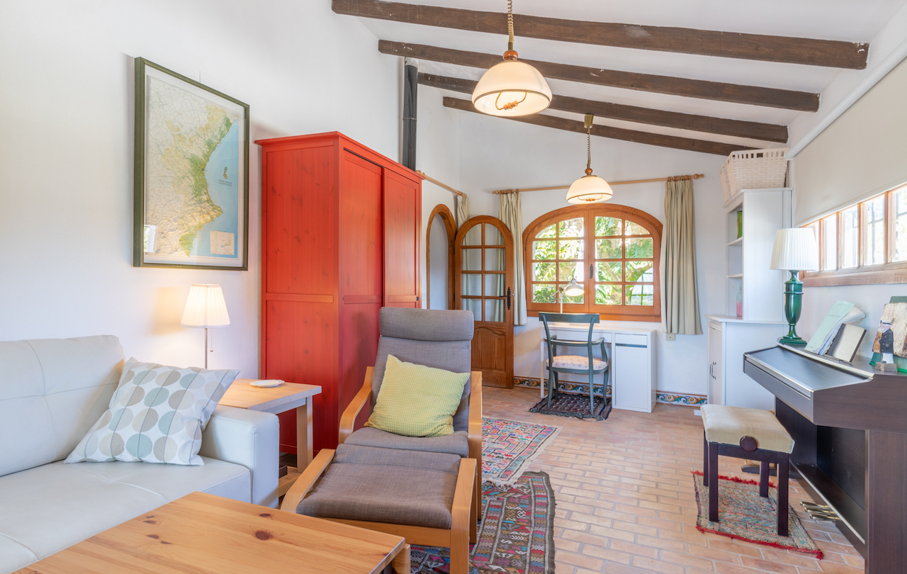 Salon avec poutres apparentes, sol en terre cuite et armoire rouge. L'espace dispose d'un coin bureau sous une fenêtre cintrée et d'un piano numérique.