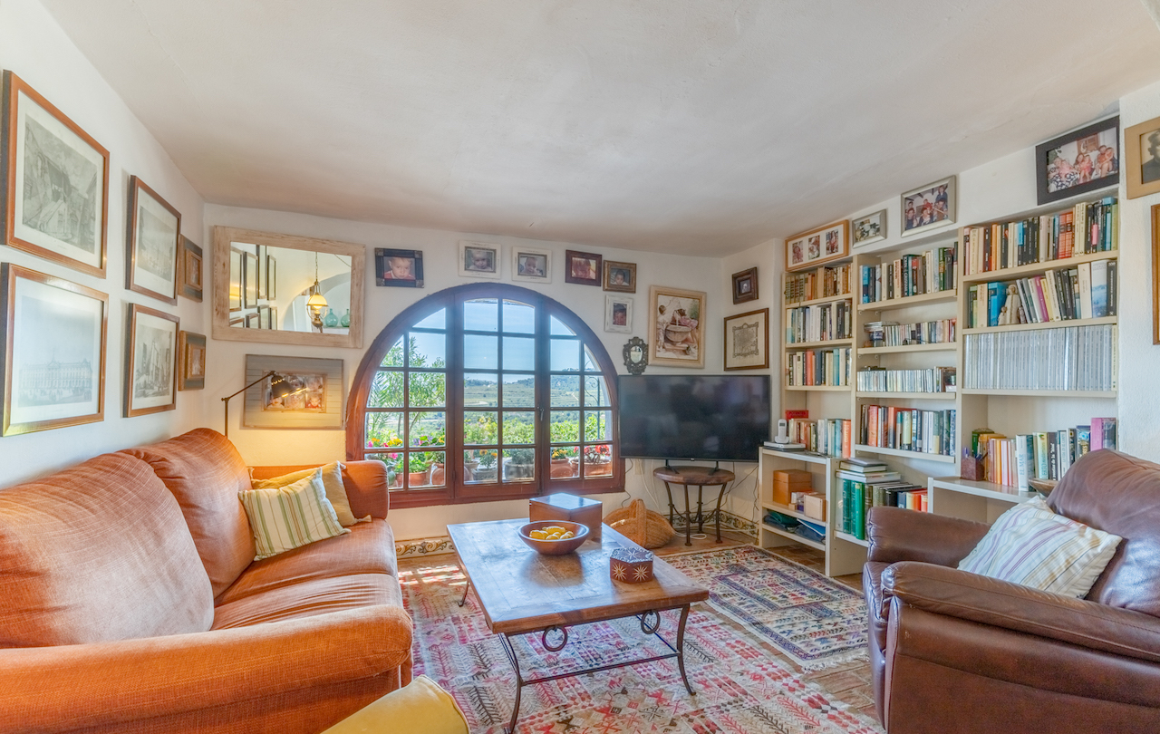 Salon bibliothèque avec une grande fenêtre cintrée en bois, étagères intégrées, sol en terre cuite avec tapis et vue dégagée sur les vignobles environnants.