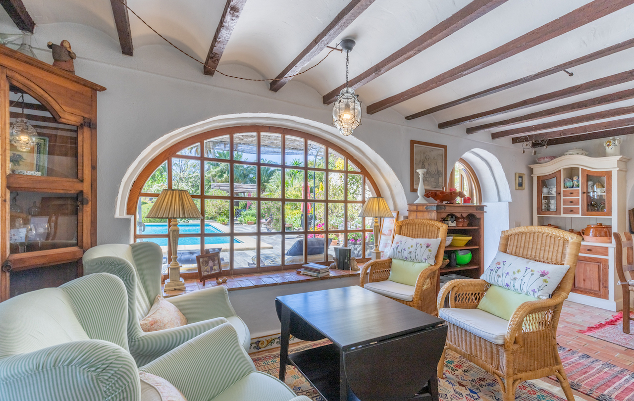 Salon de style traditionnel avec poutres apparentes, sol en terre cuite et une grande fenêtre en arche offrant une vue sur la piscine et le jardin méditerranéen.