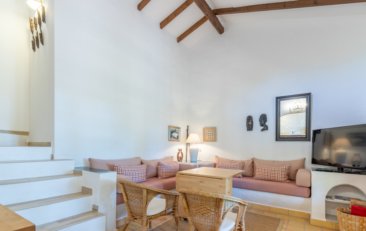 Salon rustique avec de hauts plafonds et des poutres en bois apparentes. Il comprend des banquettes intégrées, des fauteuils en osier et un sol en terre cuite traditionnelle.