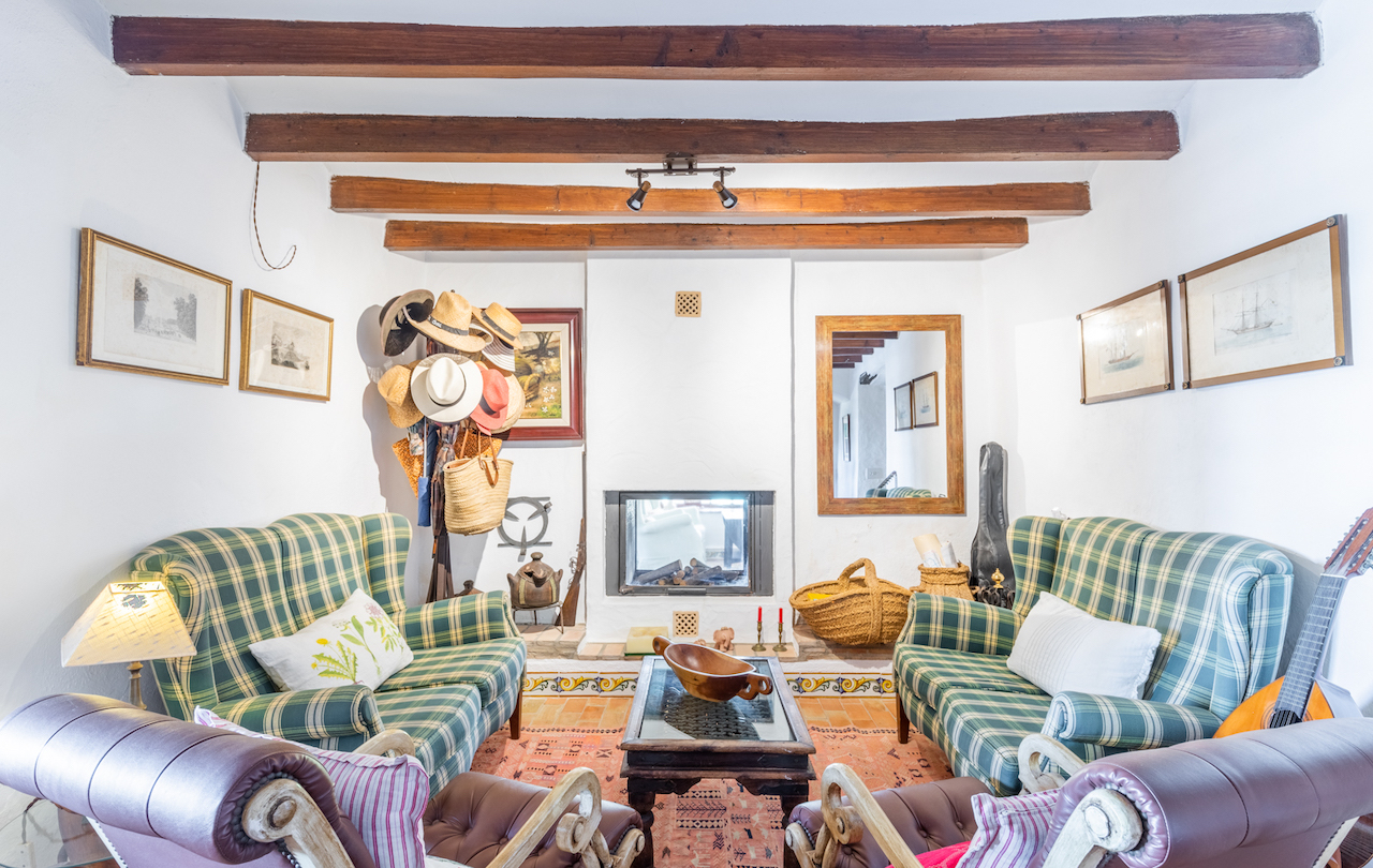 Salon rustique avec poutres apparentes en bois sombre, cheminée encastrée et sol en terre cuite. L'espace comprend des fauteuils à carreaux et une décoration traditionnelle.
