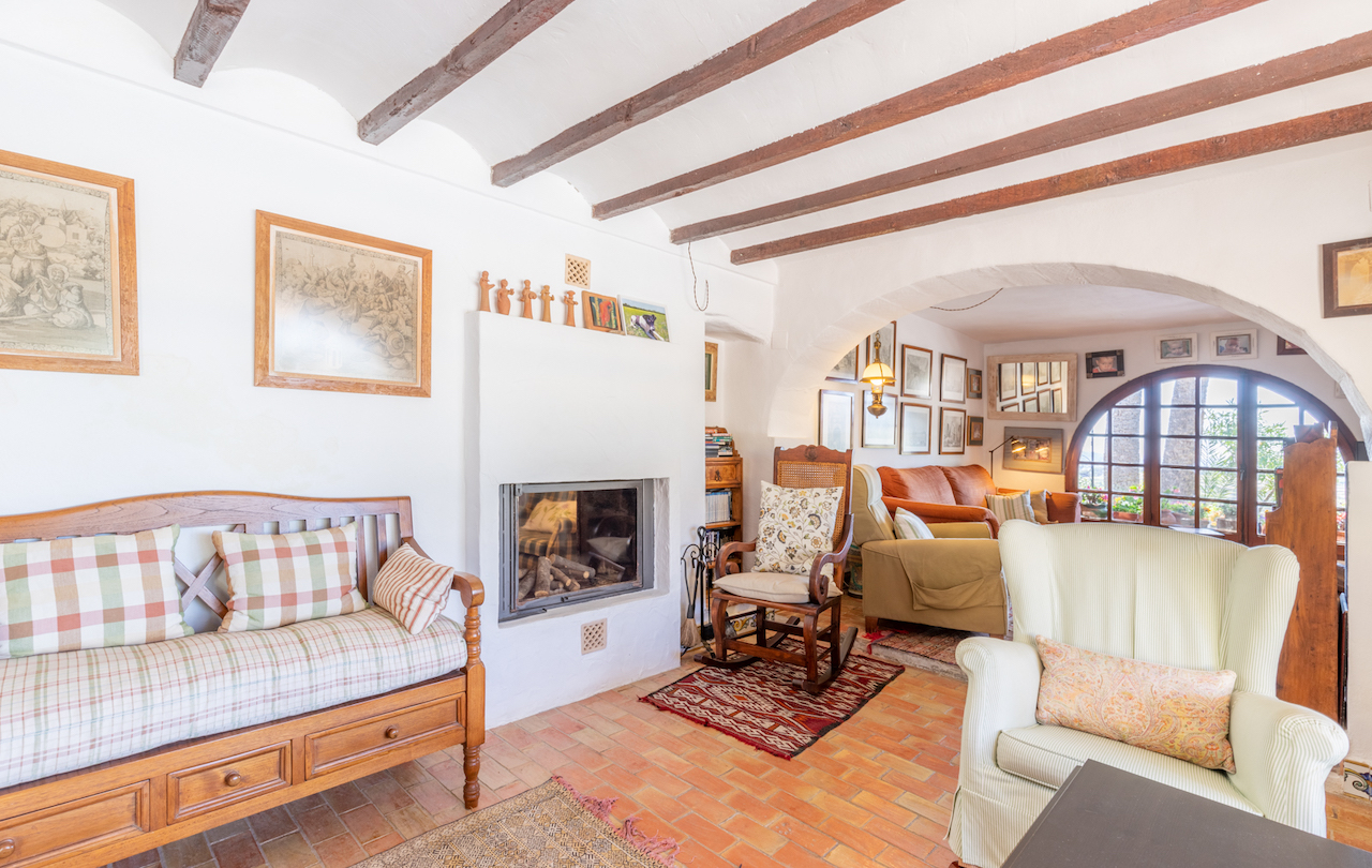 Salon traditionnel avec poutres en bois apparentes, sol en terre cuite et cheminée encastrée. Une arche mène à un espace de détente lumineux avec une fenêtre en plein cintre.