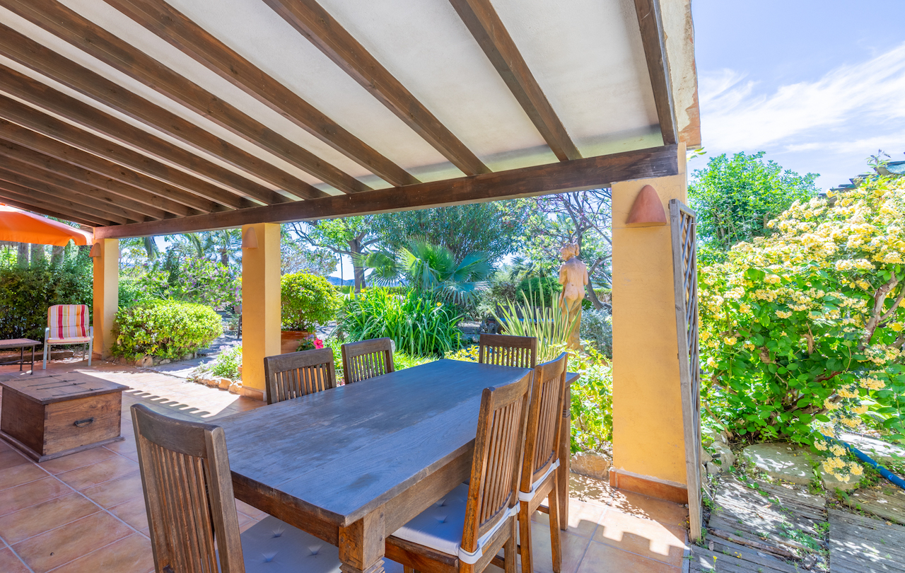 Terrasse couverte aux poutres en bois apparentes et sol en terre cuite, offrant un espace dînatoire face à un jardin méditerranéen luxuriant et fleuri.