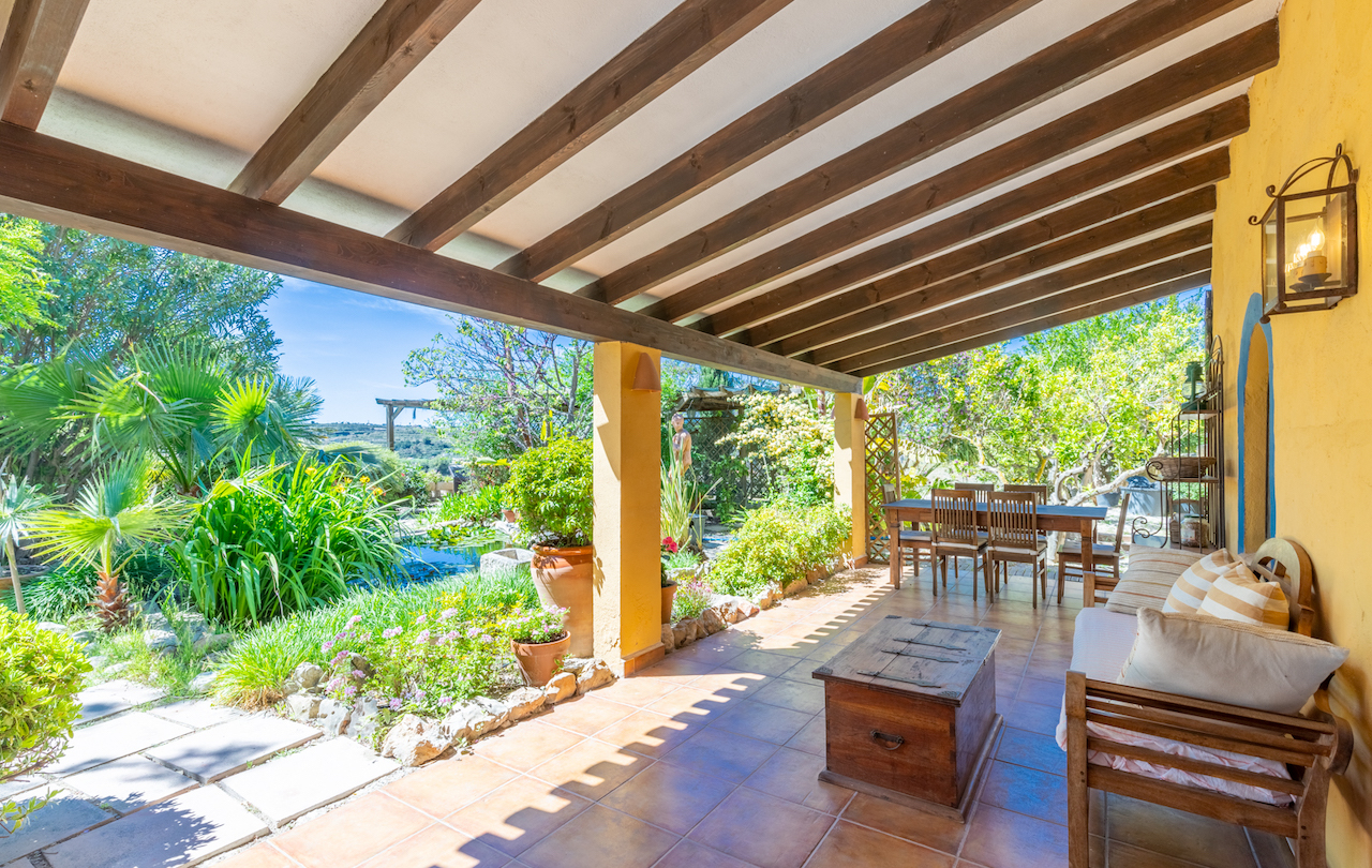Terrasse couverte avec poutres en bois apparentes, sol en terre cuite et espaces de vie extérieurs donnant sur un jardin méditerranéen avec bassin.