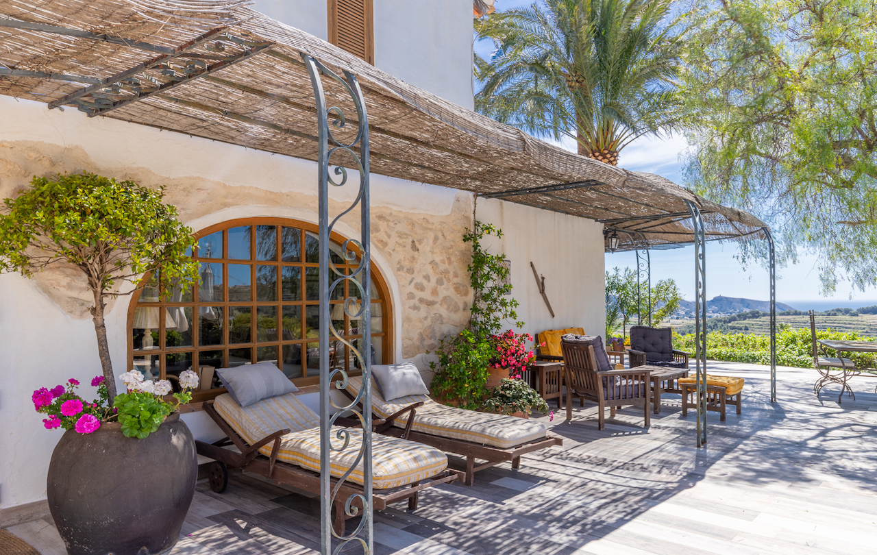 Terrasse extérieure avec pergola en canisse et fer forgé, espace détente avec bains de soleil devant une fenêtre cintrée et vue dégagée sur la vallée.