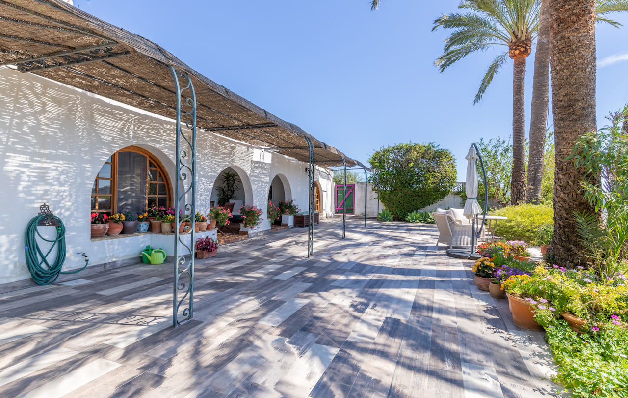 Terrasse extérieure avec sol en grès cérame aspect bois, pergola en canisse sur piliers en fer forgé et façade blanche aux arches traditionnelles fleuries.