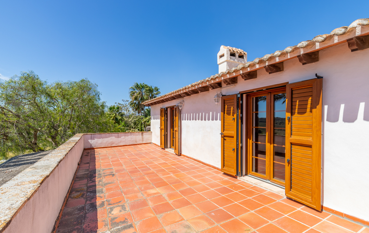 Terrasse supérieure avec carrelage en terre cuite, poutres en bois apparentes sous la toiture et portes-fenêtres en bois avec volets battants traditionnels.