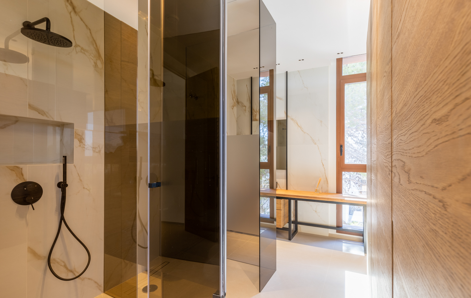 Détail de douche avec paroi fumée et coiffeuse en bois clair dans une salle de bain moderne avec carrelage veiné.