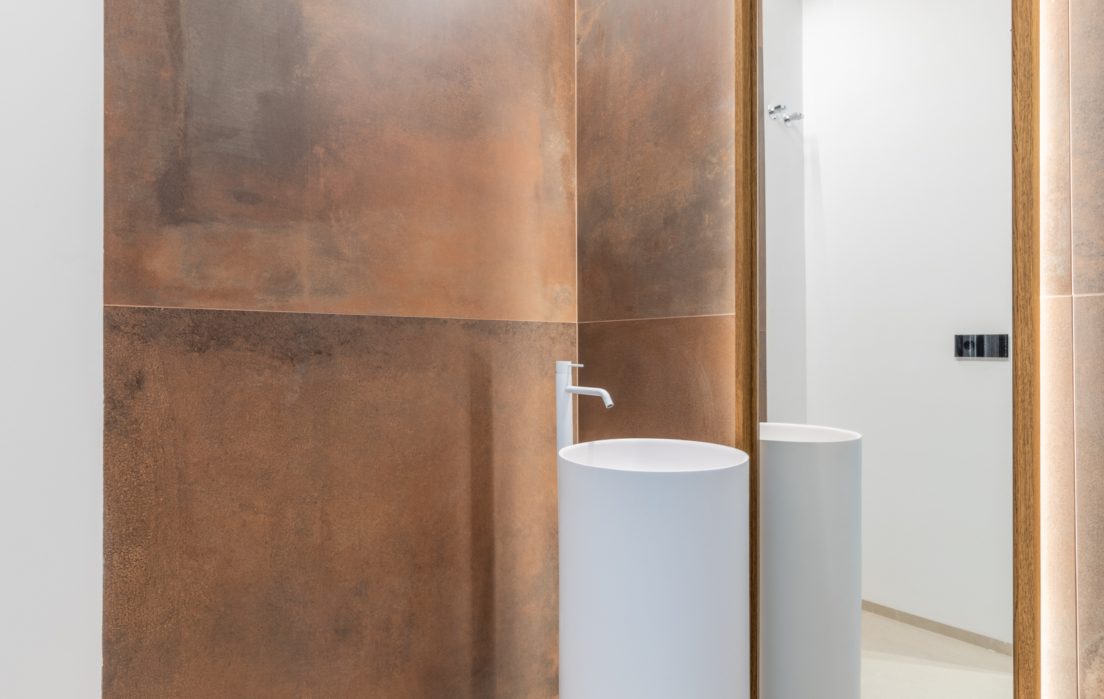 Détail de la salle de bain avec lavabo cylindrique blanc et mur carrelé couleur rouille avec miroir rétroéclairé.