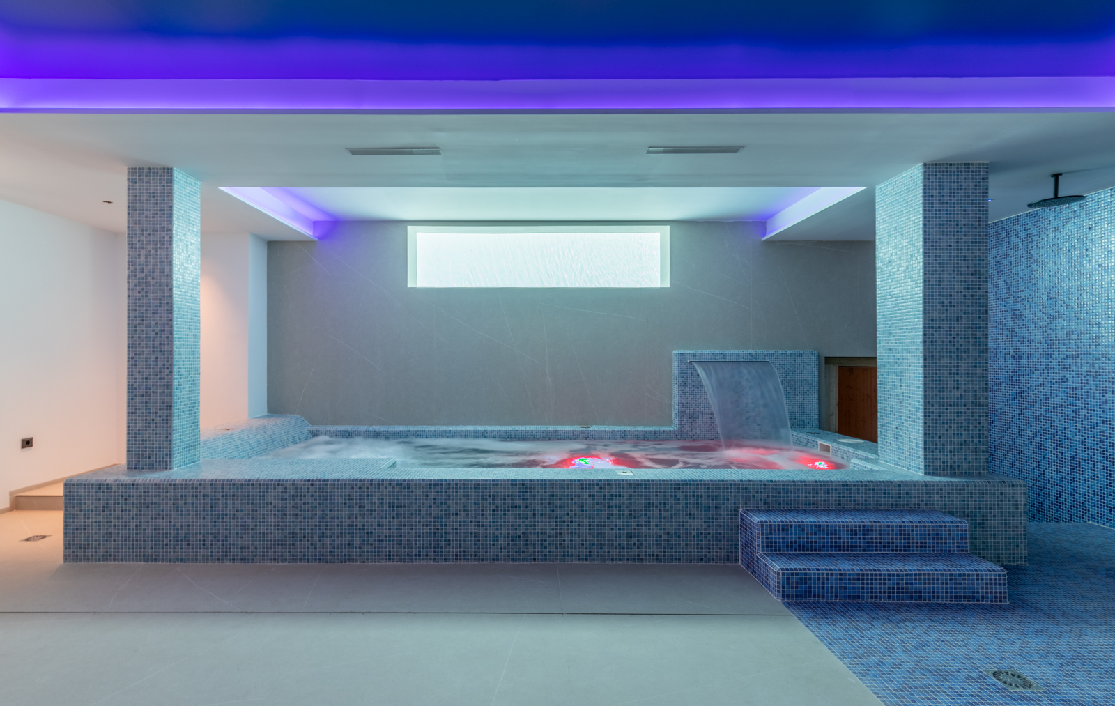 Détail du spa intérieur avec jacuzzi, cascade et douche, présentant des mosaïques bleues et un éclairage d'ambiance.
