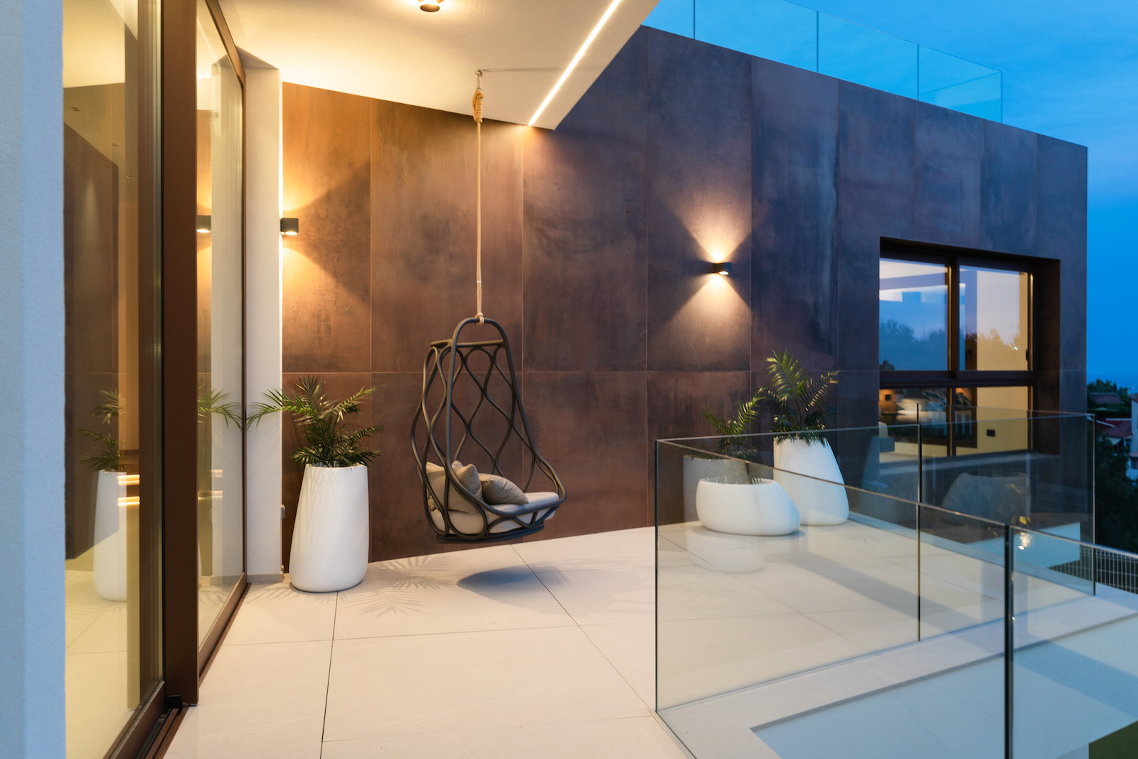 Terrasse avec mur de carreaux couleur rouille, chaise suspendue et garde-corps en verre.