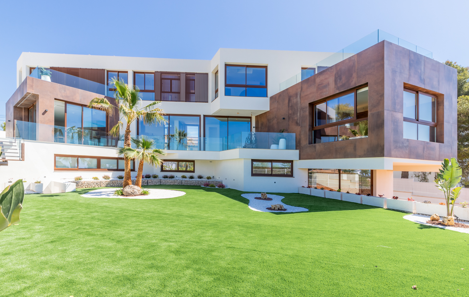 Vue extérieure de la villa avec jardin gazonné, palmiers et façade moderne avec de grandes fenêtres.