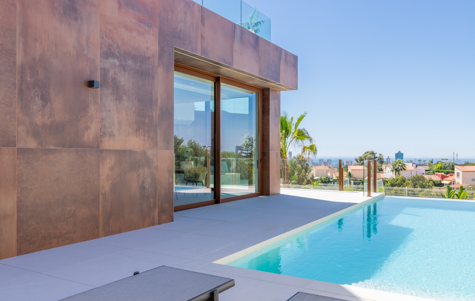 Vue sur la piscine à débordement avec façade en panneaux couleur rouille et vue panoramique sur la ville.