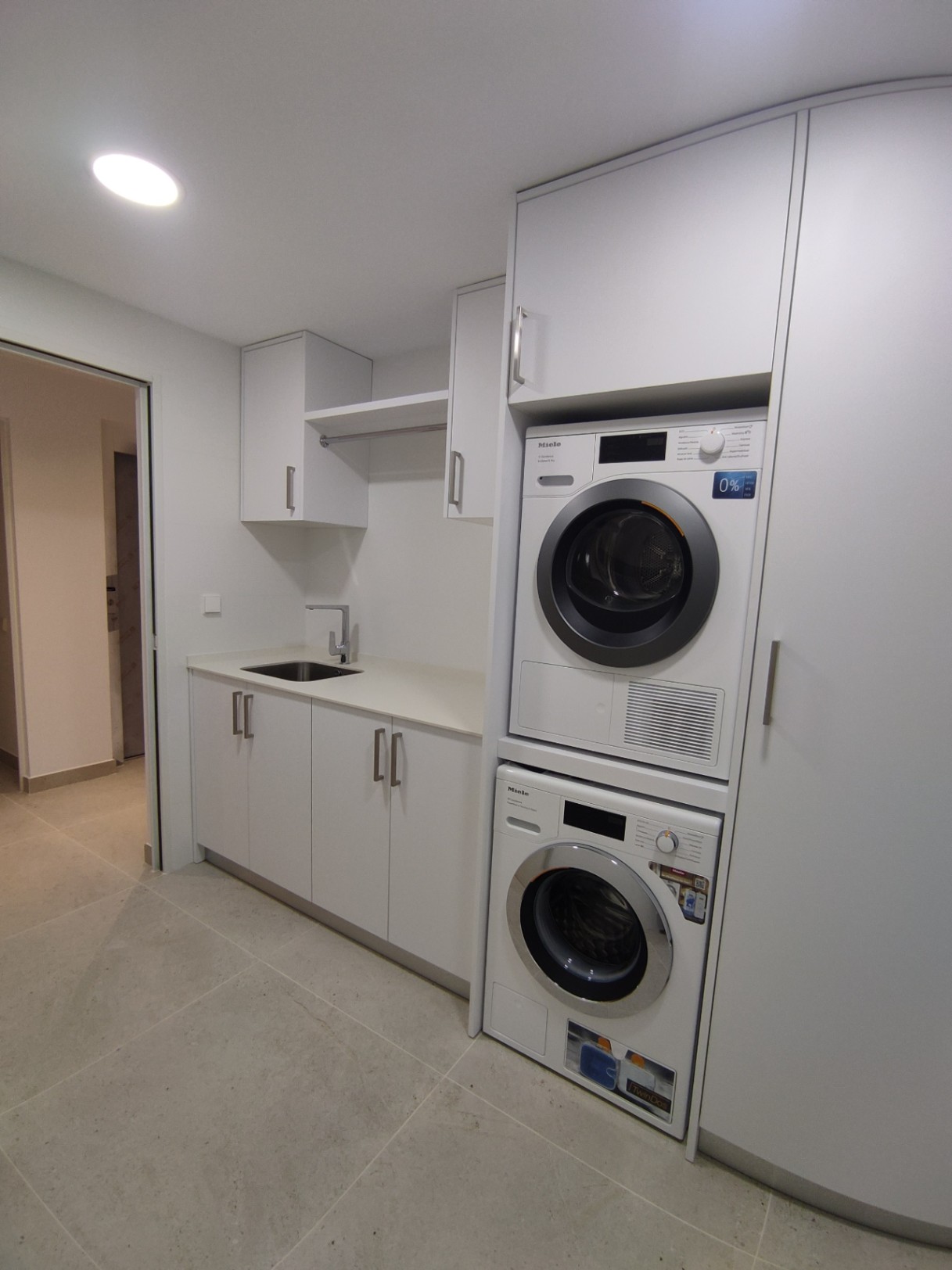 Buanderie fonctionnelle avec appareils Miele superposés, armoires blanches minimalistes, évier intégré et carrelage de sol grand format aux tons neutres.