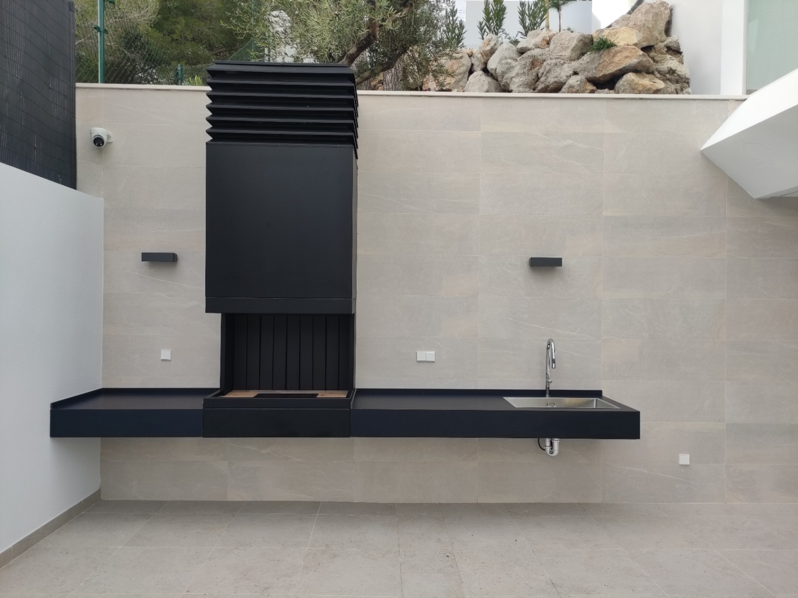 Cuisine d'été minimaliste avec barbecue en métal noir, évier intégré et plan de travail suspendu sur un mur en pierre naturelle aux tons clairs.