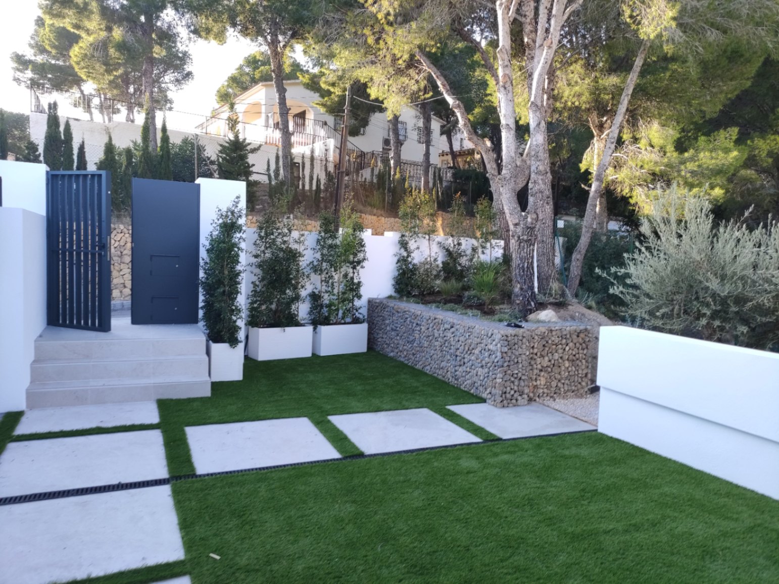 Entrée extérieure avec portail métallique moderne, marches en pierre et jardin paysager comprenant du gazon synthétique, des dalles en béton et un mur de gabions.