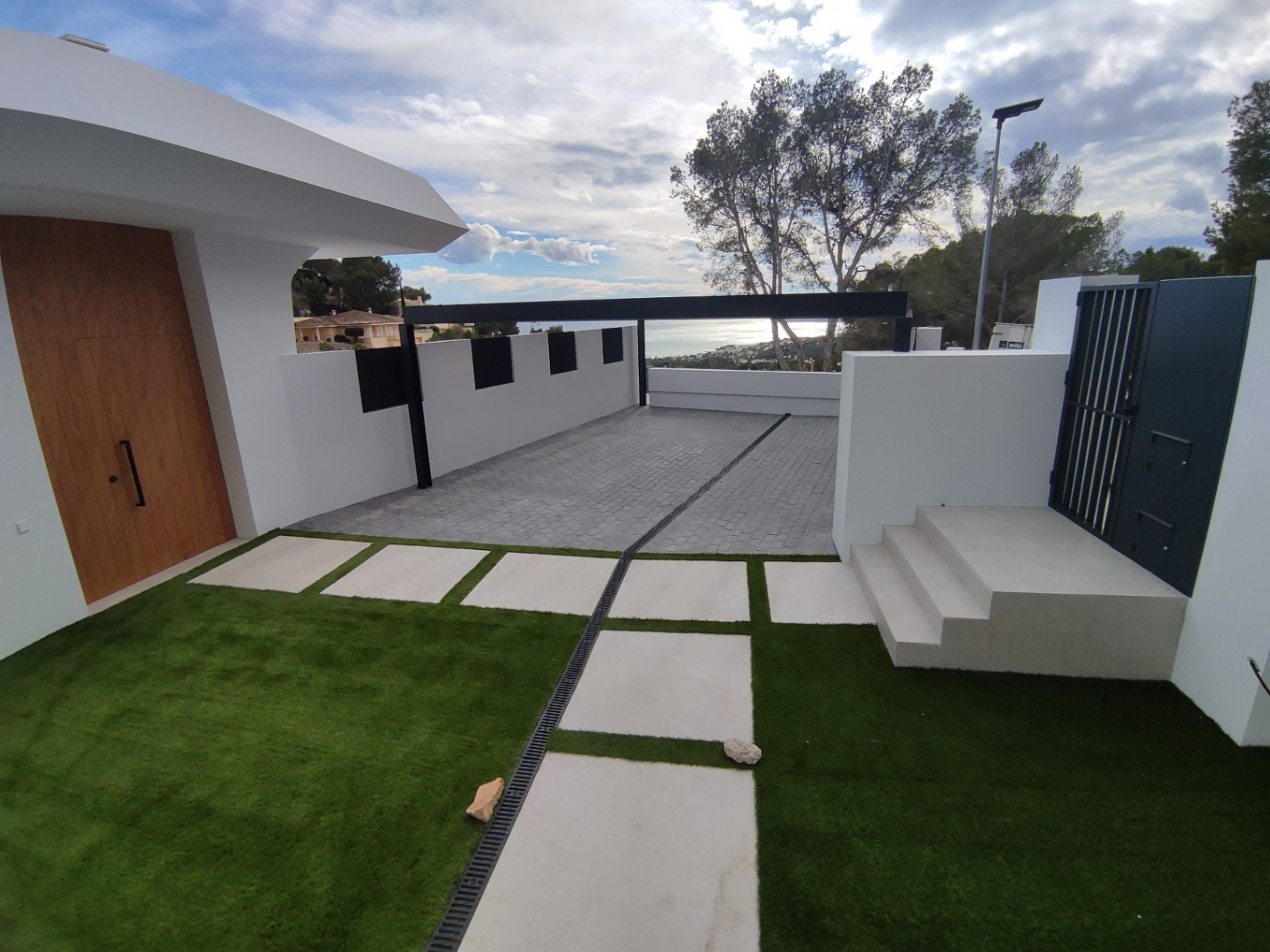 Entrée extérieure avec porte en bois, allée pavée et gazon synthétique agrémenté de dalles blanches. L'architecture moderne offre un aperçu sur l'horizon marin.