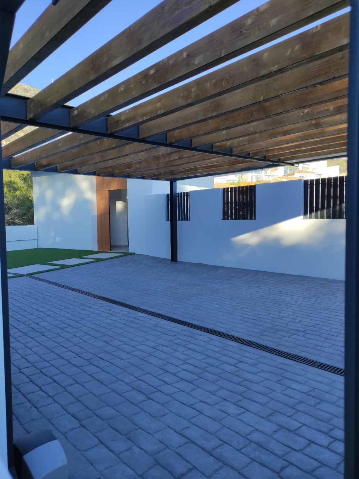 Espace de stationnement extérieur avec pergola en poutres de bois, pavage en pierre grise et accès à la villa souligné par des finitions en bois naturel.
