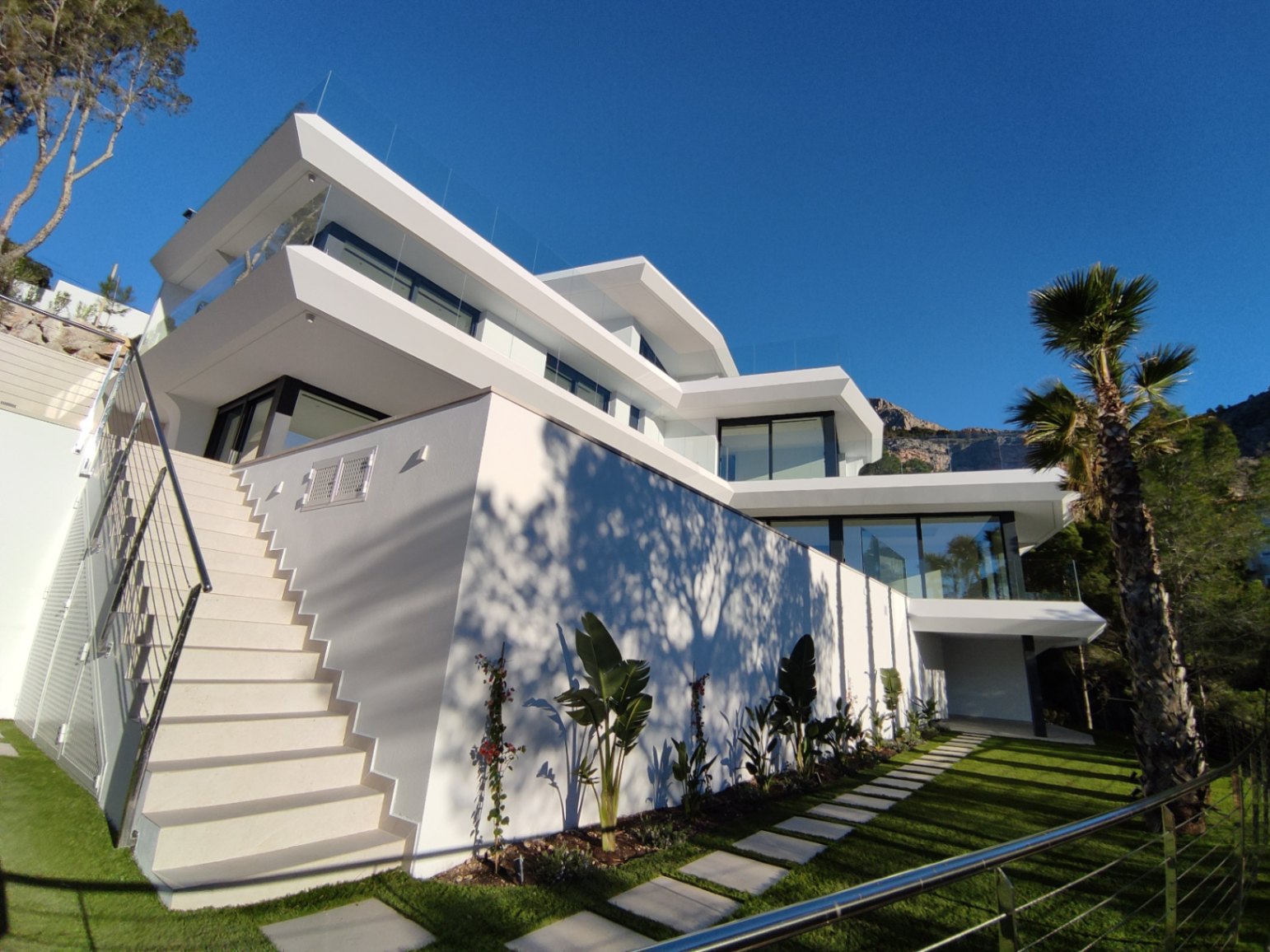 Façade contemporaine sur plusieurs niveaux avec terrasses vitrées, escalier extérieur et jardin paysager doté d'un chemin de dalles et de palmiers.
