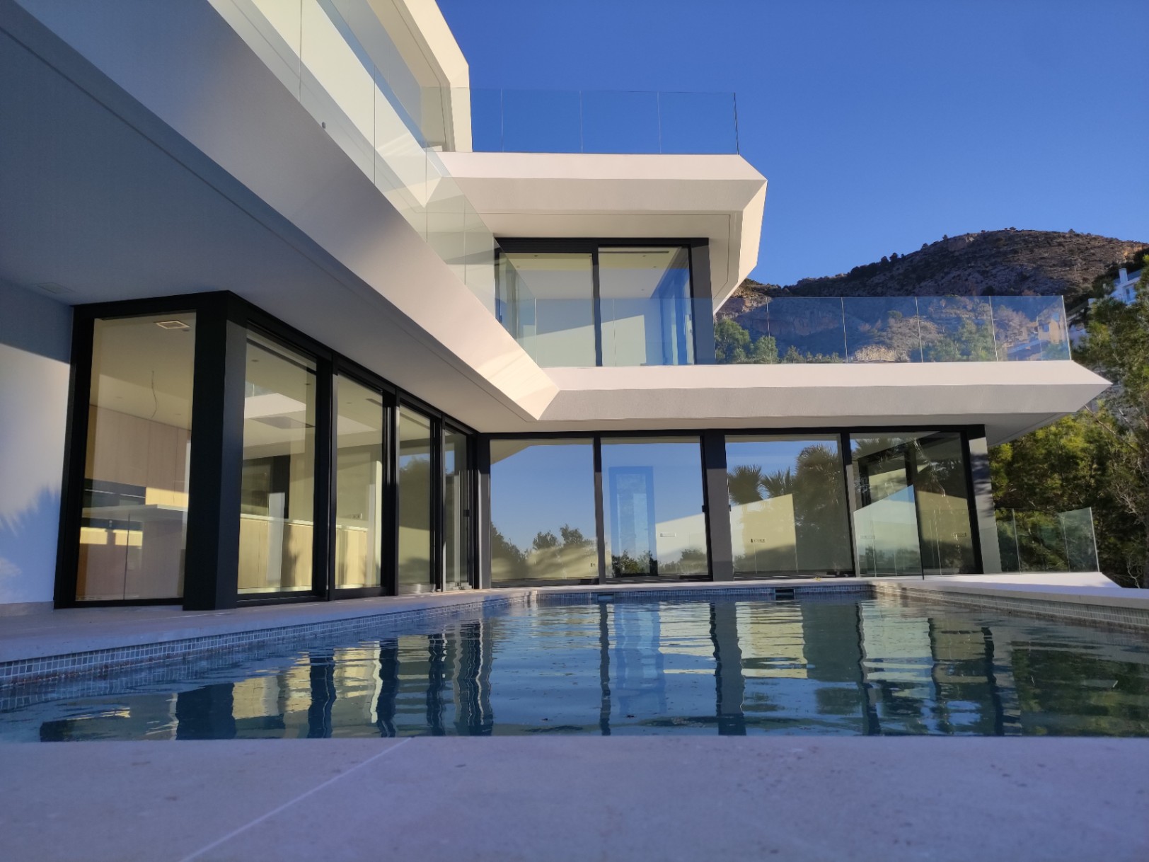Façade moderne avec de larges baies vitrées à cadre noir et une piscine à débordement reflétant l'architecture géométrique sous le ciel d'Altea Hills.