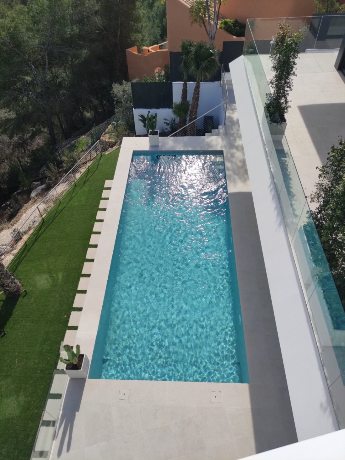 Piscine rectangulaire aux eaux cristallines, entourée d'une terrasse en pierre claire, d'un espace en gazon synthétique et de garde-corps en verre épurés.