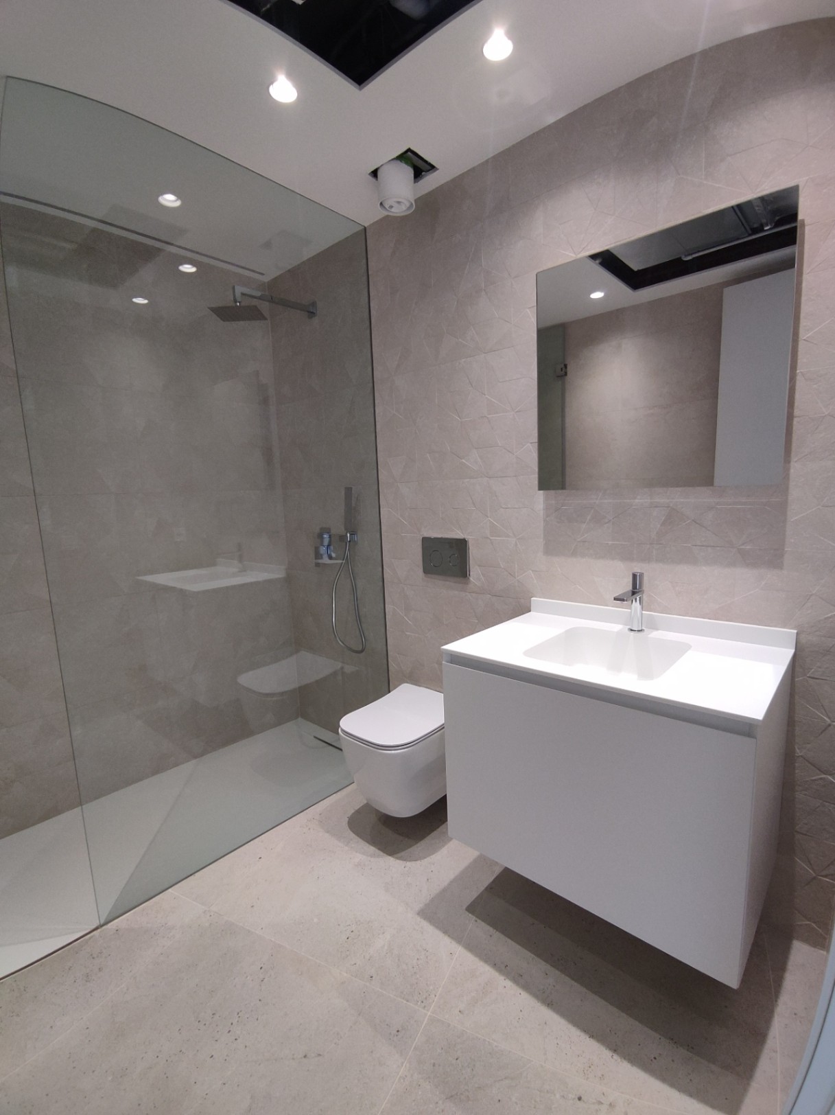 Salle de bains moderne avec douche à l'italienne vitrée, toilettes suspendues et meuble vasque flottant blanc sur carrelage mural à relief géométrique.
