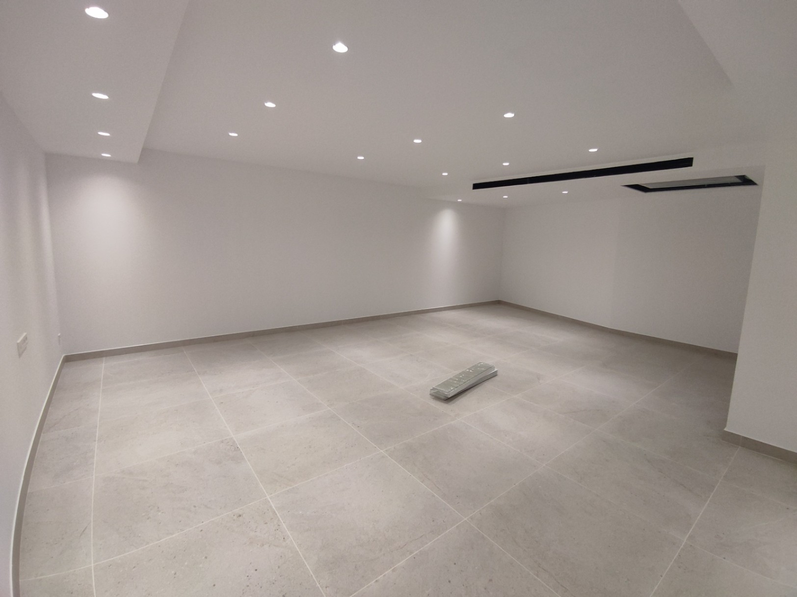 Vaste pièce polyvalente avec revêtement de sol en grès cérame grand format, murs blancs et éclairage par spots LED encastrés au plafond.