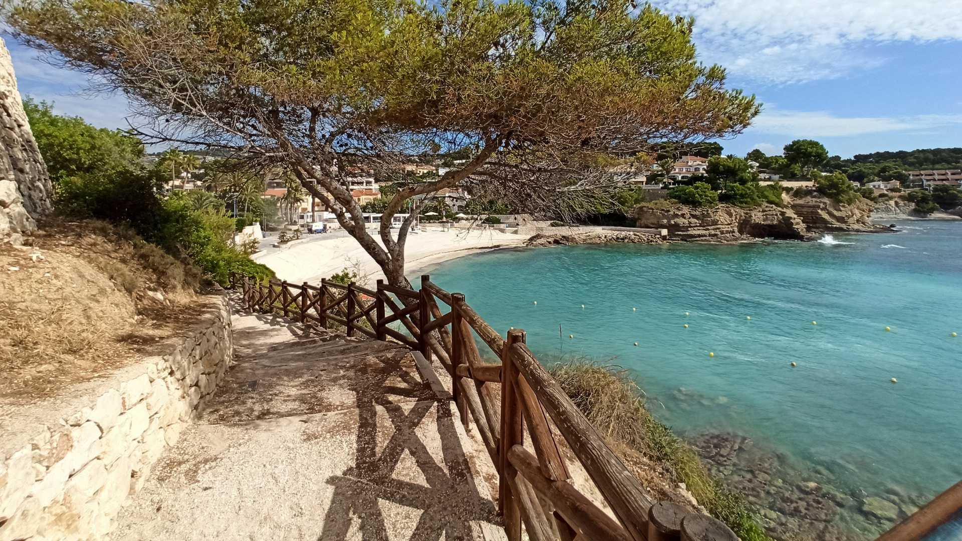 Sentier côtier avec garde-corps en bois et murs en pierre naturelle, menant directement à la plage de sable fin de La Fustera et ses eaux turquoise.