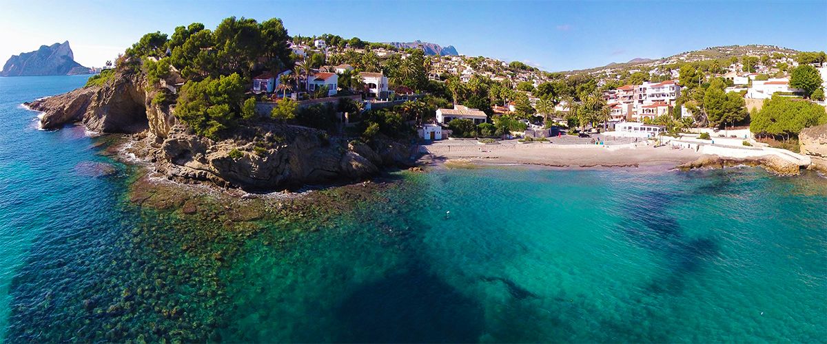 Vue aérienne de la plage de La Fustera avec ses eaux turquoises, ses falaises rocheuses et le Peñón de Ifach au loin, entourée de pins et de villas côtières.