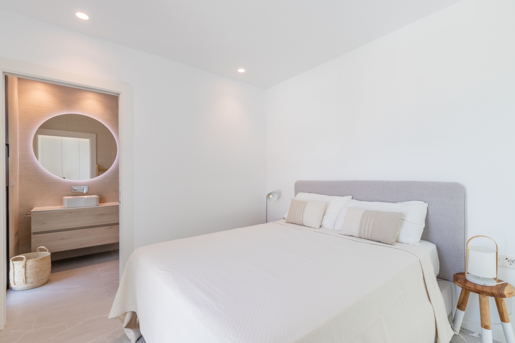 Chambre lumineuse avec salle de bain attenante. Lit double avec tête de lit grise et linge blanc. Miroir circulaire lumineux dans la salle de bain.