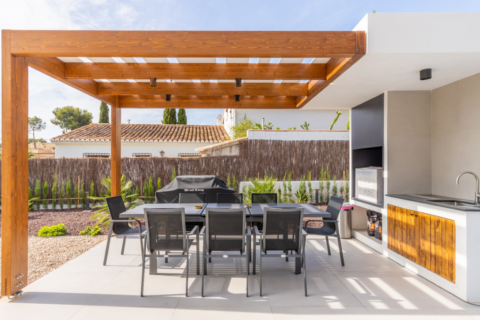 Détail de la salle à manger extérieure avec une pergola en bois et une cuisine extérieure avec évier et barbecue.