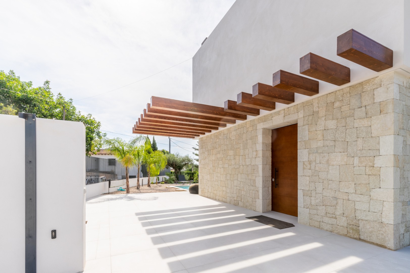 Entrée principale avec mur en pierre et pergola en bois projetant des ombres sur le sol.