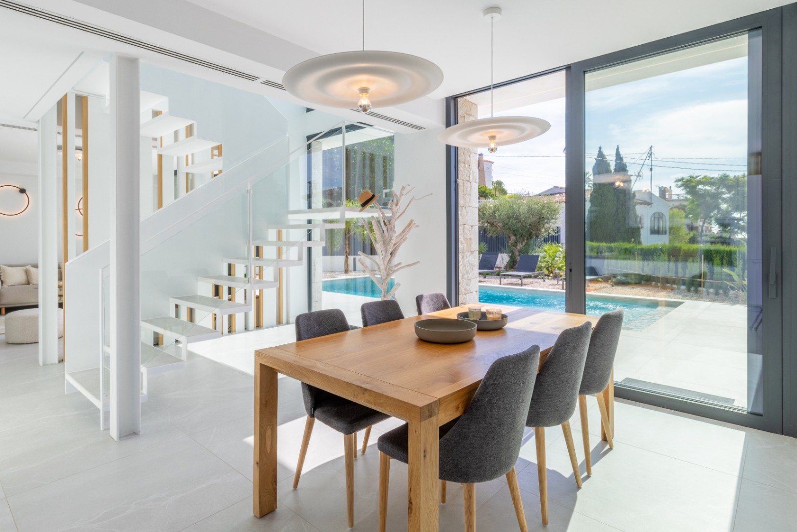 Salle à manger lumineuse avec table en bois, vue sur la piscine et escalier flottant blanc.