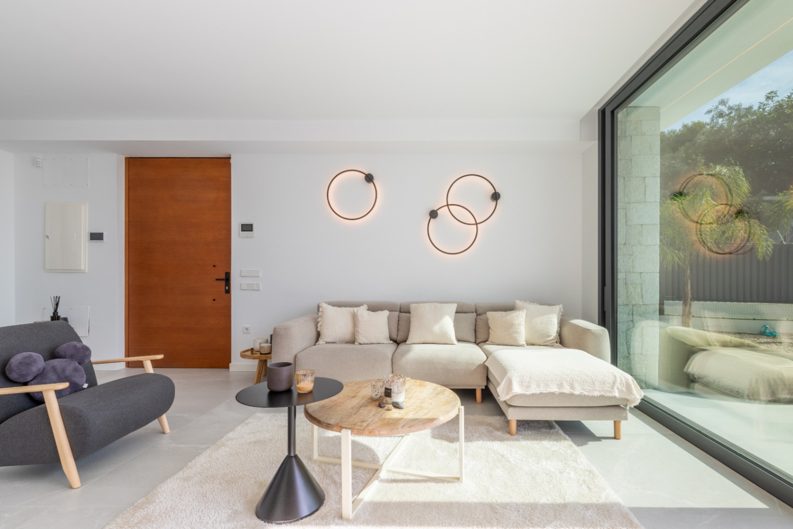 Salon lumineux avec canapé beige, tapis à poils longs et lampes circulaires. Porte en bois et grande fenêtre avec vue extérieure.