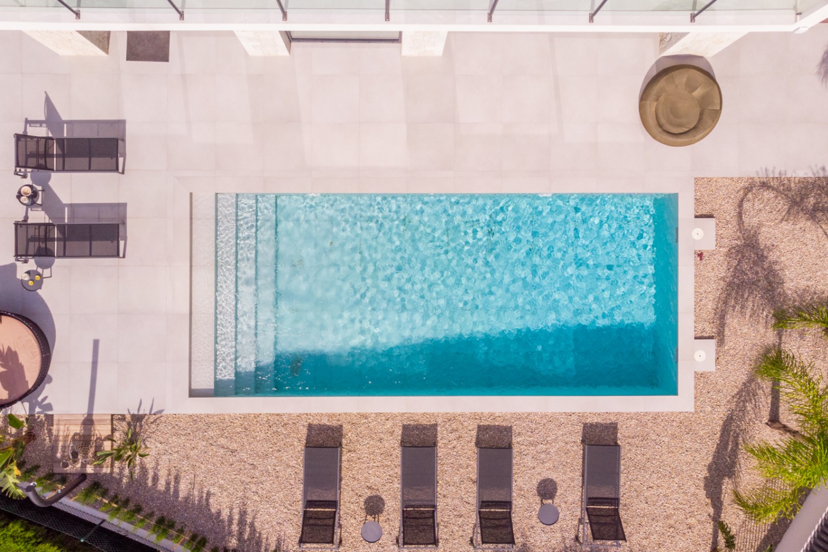 Vue aérienne de la piscine rectangulaire avec marches, entourée de carreaux clairs et de gravier. Des chaises longues et de la végétation complètent l'ensemble.