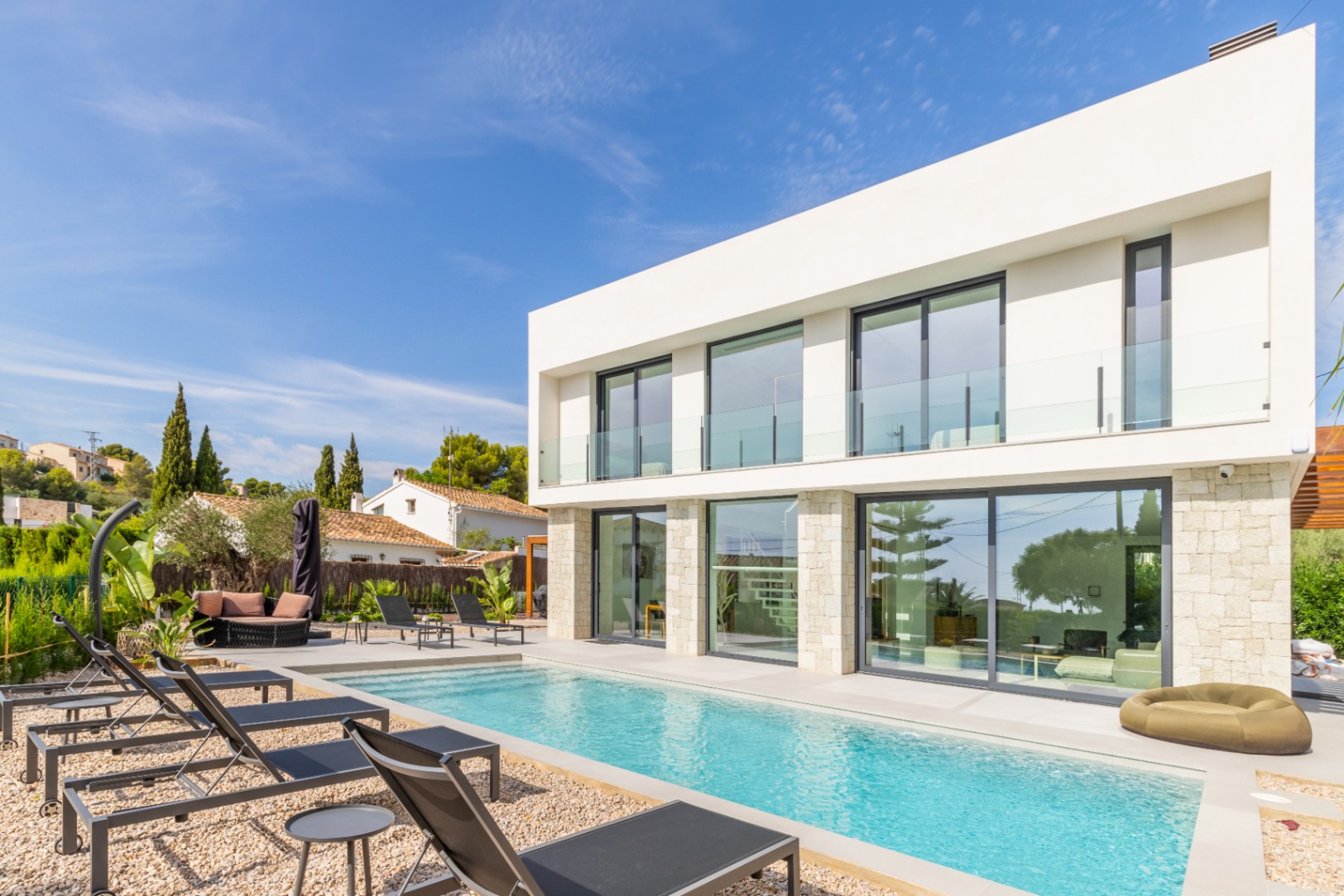 Vue extérieure de la villa avec piscine allongée, entourée de gravier et de chaises longues noires.