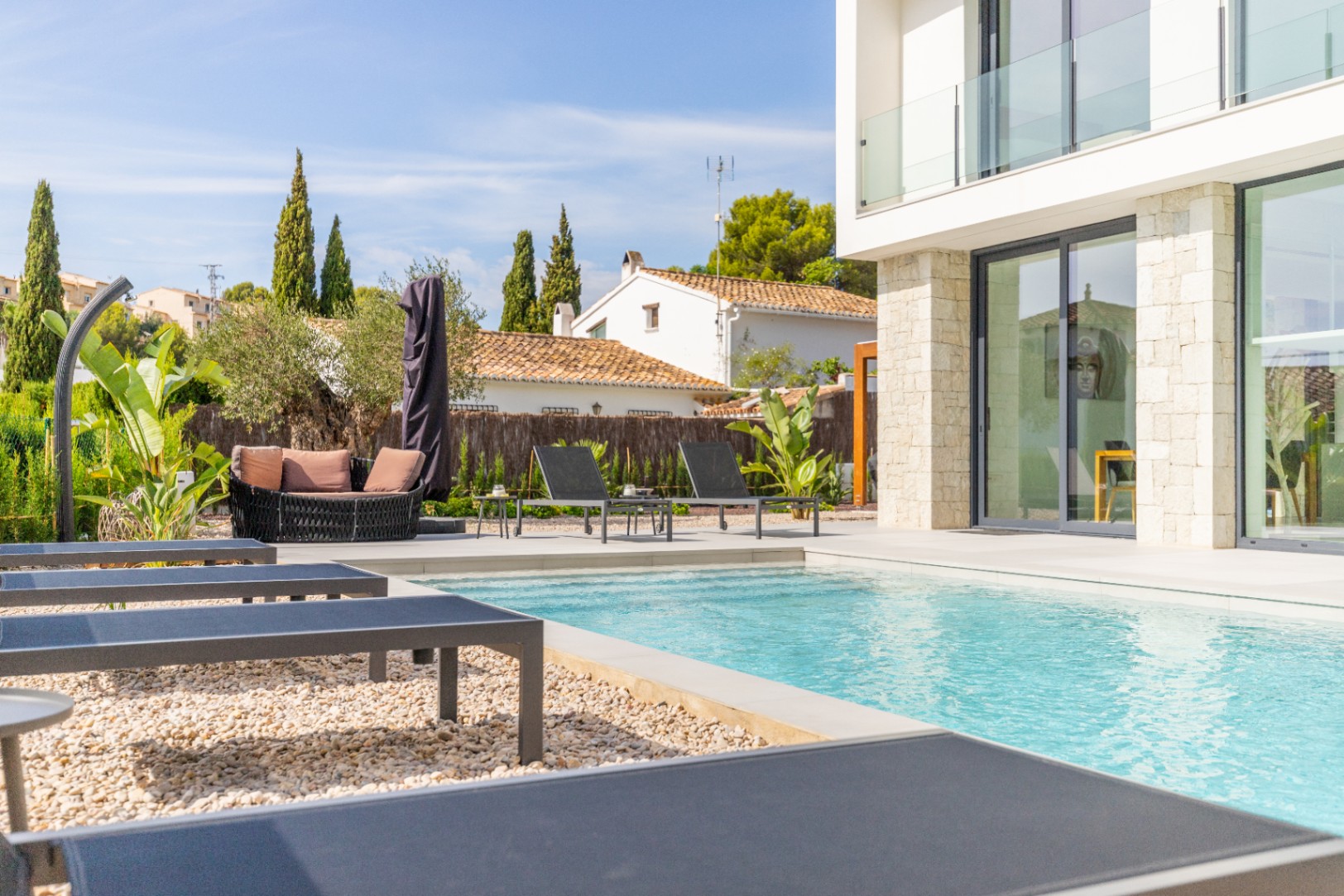Vue sur la piscine avec chaises longues grises et coin salon extérieur avec coussins couleur terre cuite.