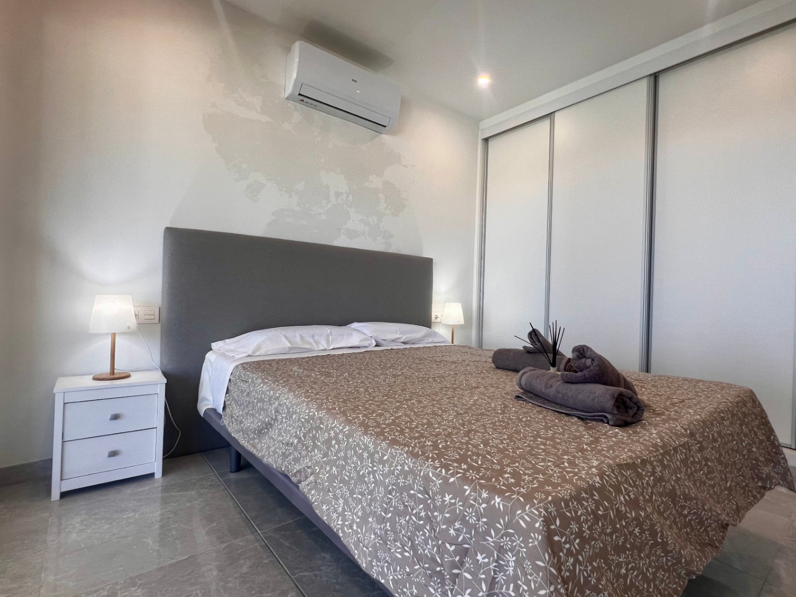 Chambre contemporaine avec dressing à portes coulissantes, climatisation réversible et tête de lit rembourrée sur un sol en carrelage gris.
