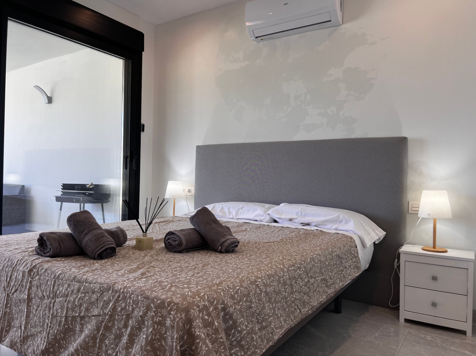 Chambre contemporaine avec tête de lit en tissu gris, climatisation split et accès direct à la terrasse par de grandes baies vitrées aux cadres noirs.