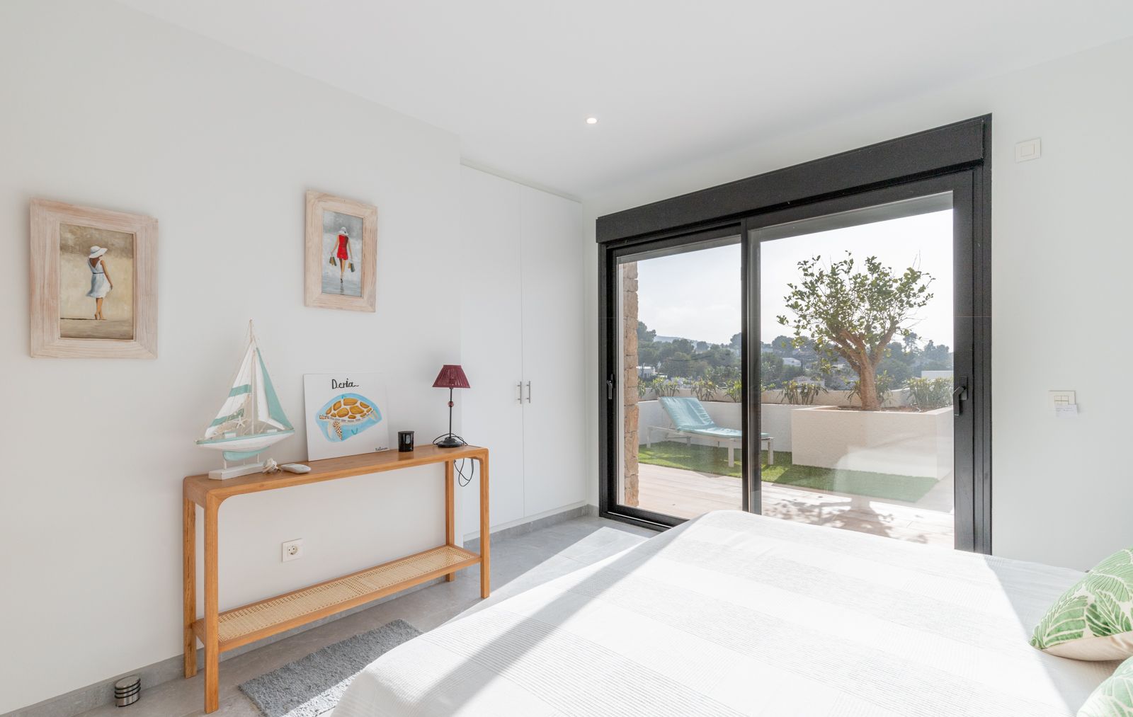 Chambre lumineuse avec placards intégrés blancs et baie vitrée ouvrant sur une terrasse privée avec gazon synthétique et transat.