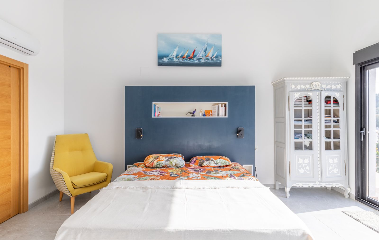Chambre moderne avec tête de lit bleue intégrée, fauteuil jaune et armoire classique. Comprend la climatisation et un accès direct vers l'extérieur par une baie vitrée.